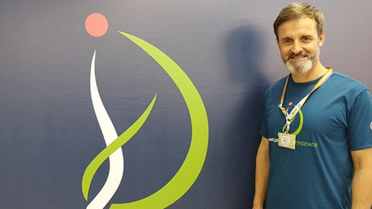 Uma pessoa vestindo uma camiseta azul está ao lado de um logotipo estilizado verde e vermelho na parede.
