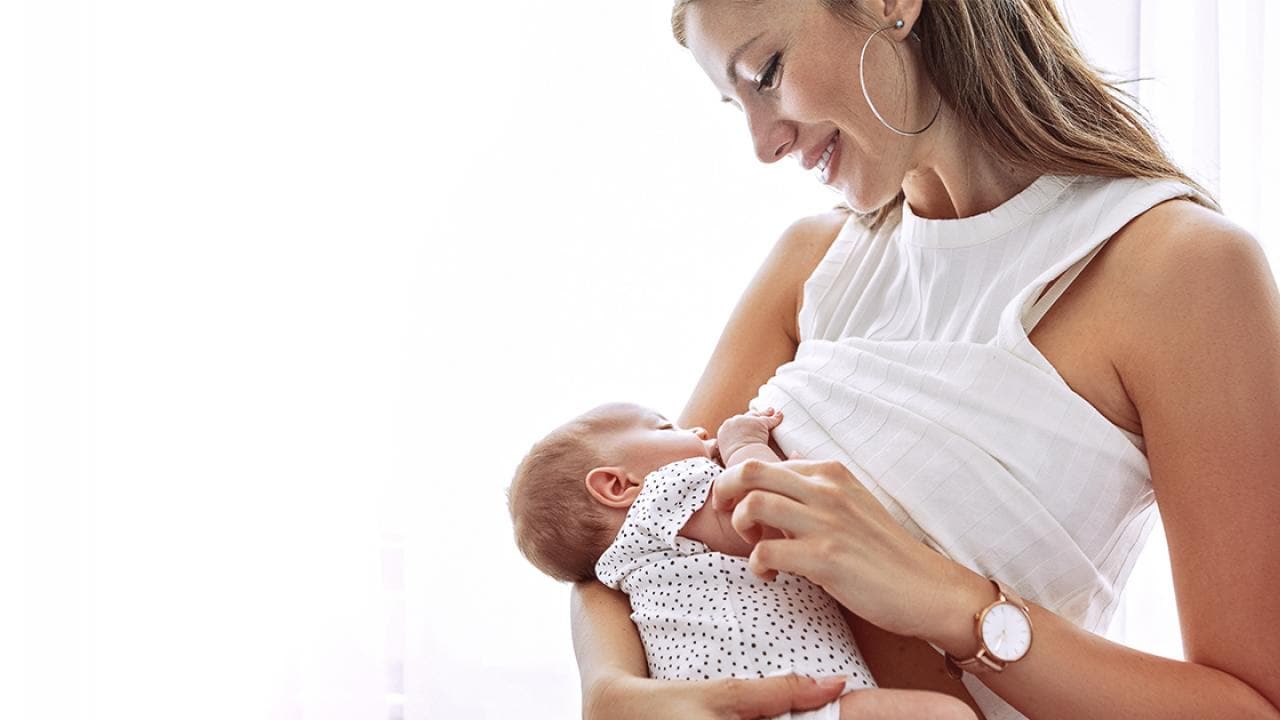 Uma mulher de vestido branco segura um bebê, olhando para ele com uma expressão gentil.