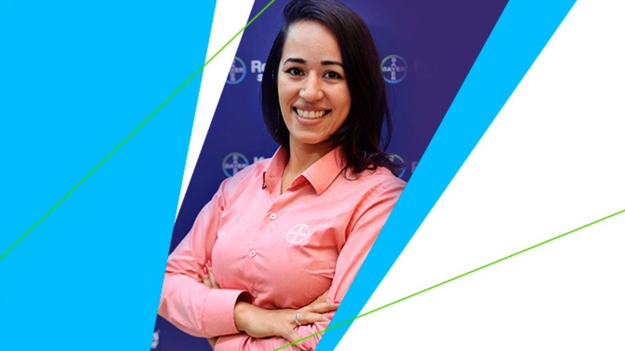 Uma pessoa de camisa rosa está sorrindo com os braços cruzados sobre um fundo azul com formas geométricas brancas e azuis.