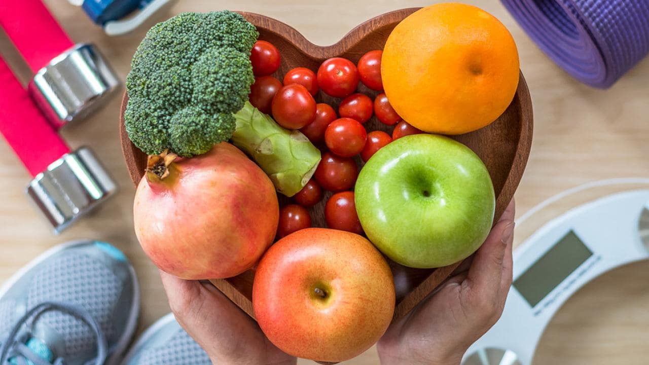 Uma tigela de madeira em formato de coração contém brócolis, tomates-cereja, uma laranja, uma romã e uma maçã verde. Perto dela, há um par de halteres, tênis, um tapete de yoga e uma balança.