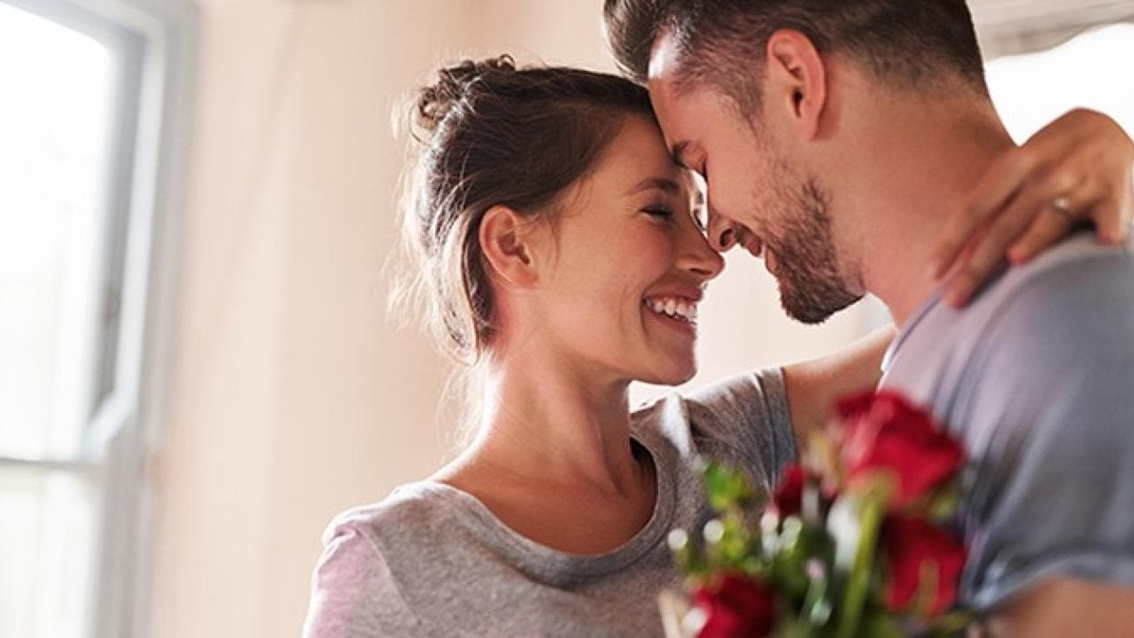 Um casal sorri e se abraça, segurando um buquê de rosas vermelhas, perto de uma janela suavemente iluminada.
