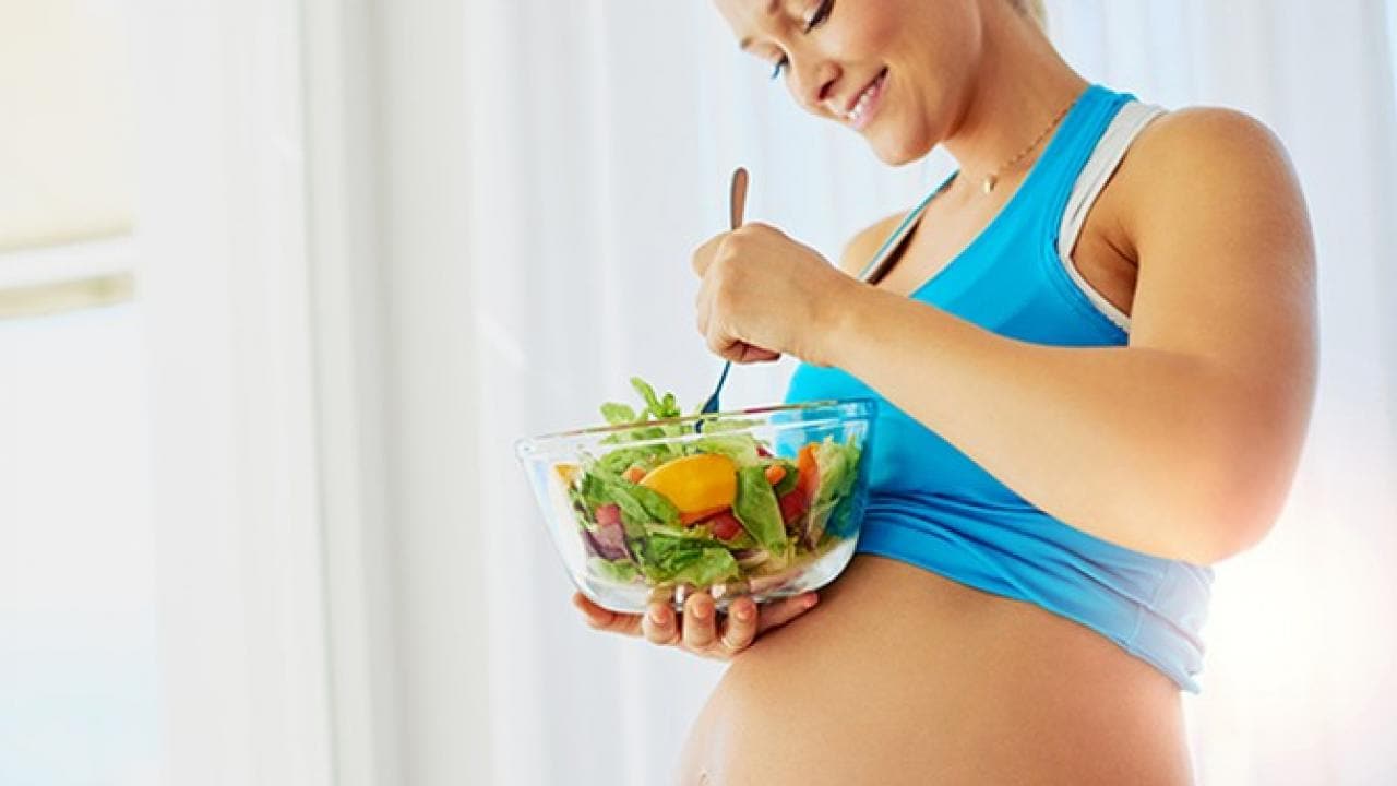 Uma mulher grávida usando um sutiã esportivo azul segura uma tigela de salada e um garfo, sorrindo enquanto olha para a comida.