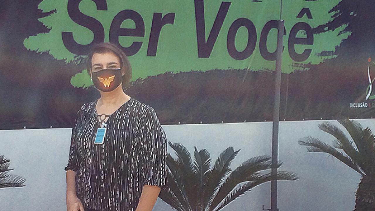 Uma pessoa está em frente a uma placa verde e preta, vestindo uma camisa estampada e uma máscara com um desenho de borboleta.