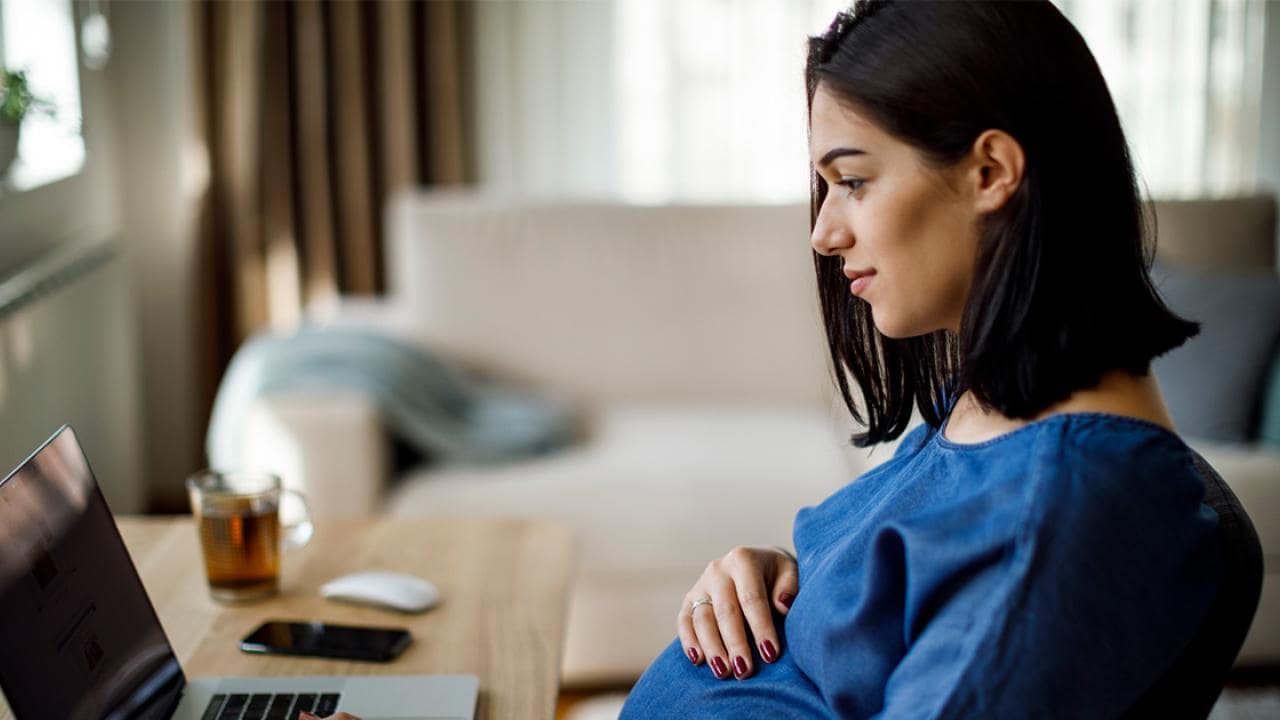 Uma mulher de blusa azul está sentada à mesa, olhando para um laptop. Ela tem a mão na barriga. Um telefone, um mouse e uma xícara estão ao lado dela, com um sofá ao fundo.