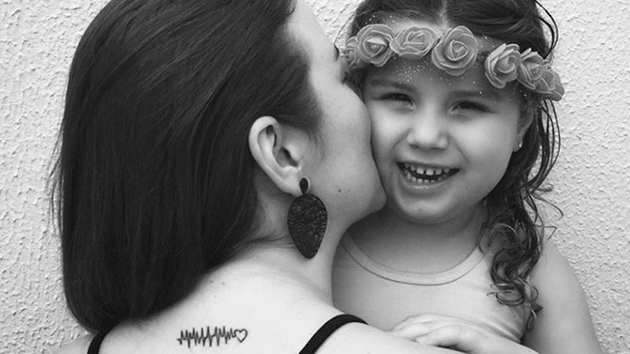 Uma mulher beija uma criança sorridente na bochecha. A criança usa uma faixa floral na cabeça. A mulher tem uma tatuagem de um padrão de batimento cardíaco com um coração no final em seu ombro. Imagem em preto e branco.