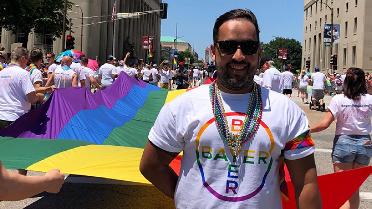 Uma pessoa vestindo uma camisa do Bayer Pride e colares de contas em frente a uma grande bandeira do arco-íris em um desfile.