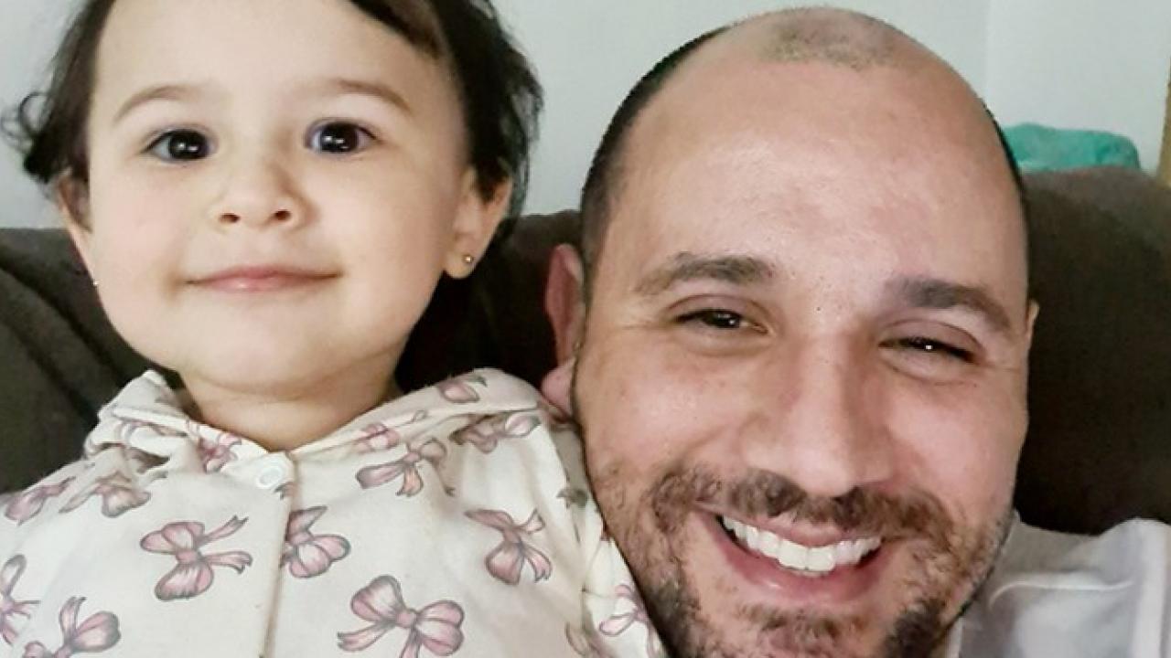 Um homem sorridente e uma criança pequena com cabelos escuros sentam-se juntos em um sofá. A criança está vestindo um moletom com capuz de cor clara com padrões de laço.
