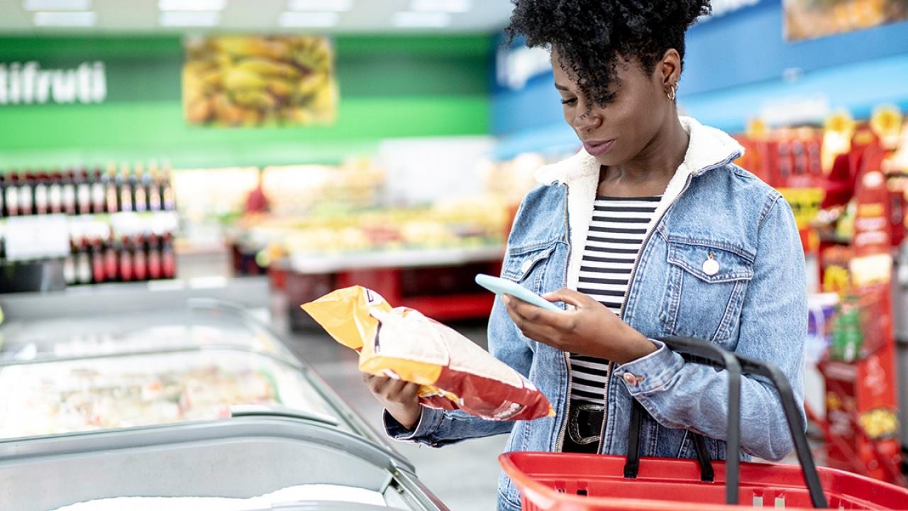 Uma pessoa vestindo uma jaqueta jeans segura uma cesta de compras e verifica um saco de batatas fritas enquanto usa um smartphone no corredor de um supermercado.