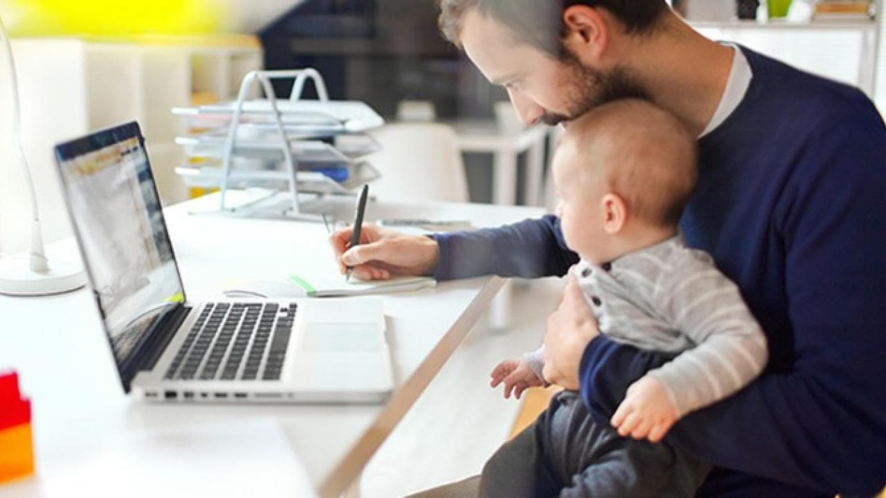 Uma pessoa trabalha em um laptop enquanto segura um bebê.