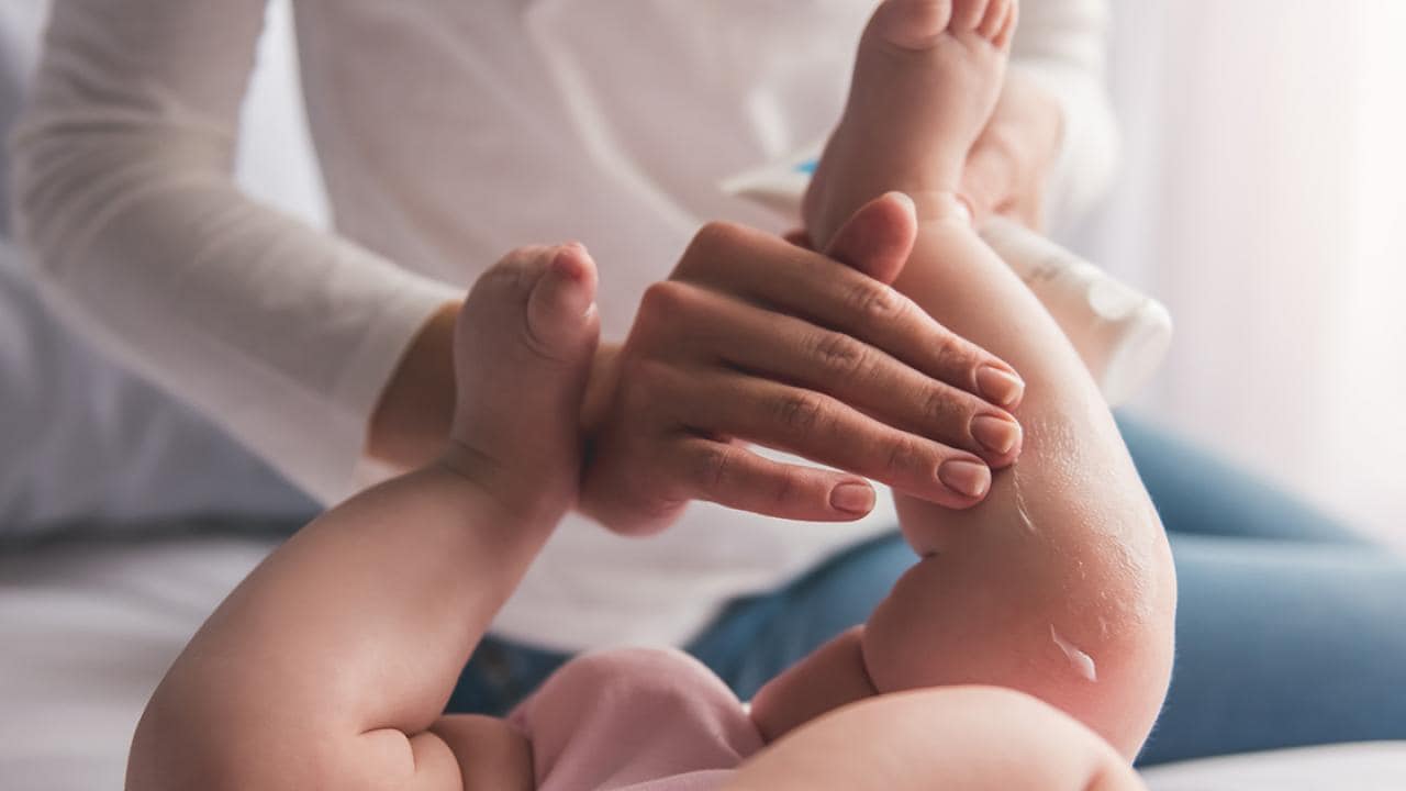 Uma pessoa aplica loção na perna de um bebê enquanto ele está deitado na cama.