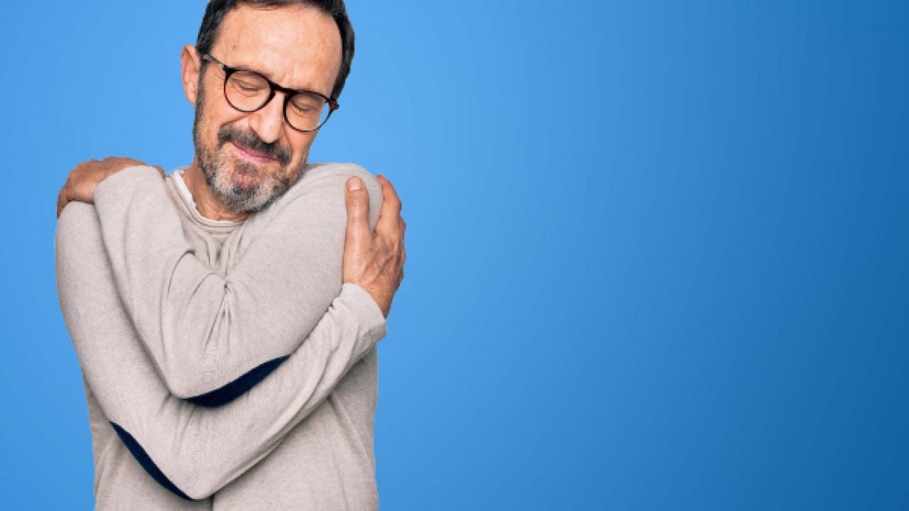 Um homem de óculos e um suéter cinza se abraça contra um fundo azul.