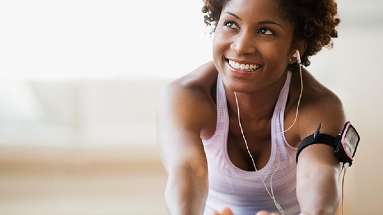 Mulher em traje de ginástica se alonga em ambientes fechados, sorrindo, com fones de ouvido e um dispositivo de braçadeira.