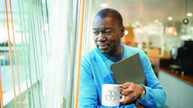 A person in a blue sweater holds a coffee mug and a folded newspaper, looking out of a window in an office setting.