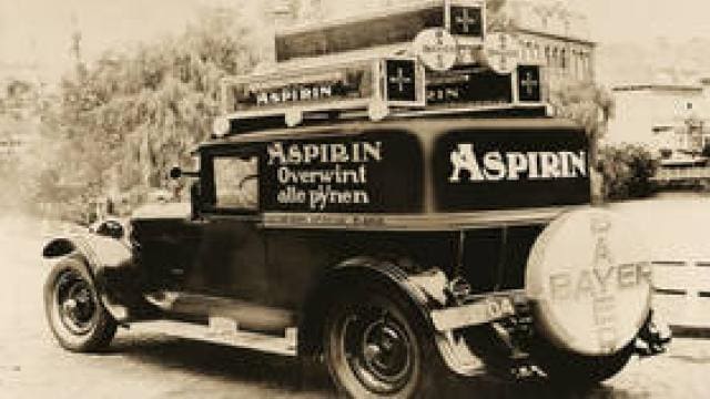 An antique car with "Aspirin" ads in German, promoting pain relief, displays a large Bayer logo on the spare tire.