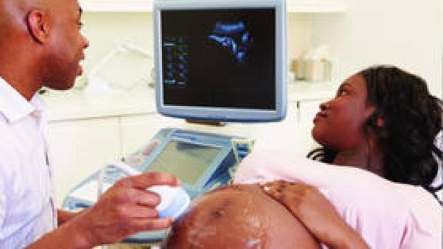 A doctor performs an ultrasound on a pregnant woman who is lying down. The ultrasound image is visible on a monitor.