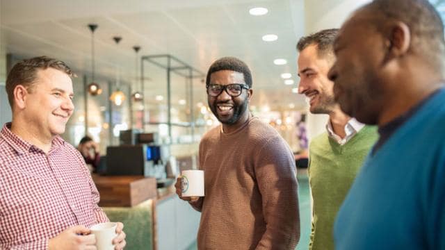 Four men are standing and smiling while holding mugs in an indoor setting.