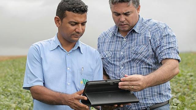 Two men stand in a field reviewing information on a tablet.