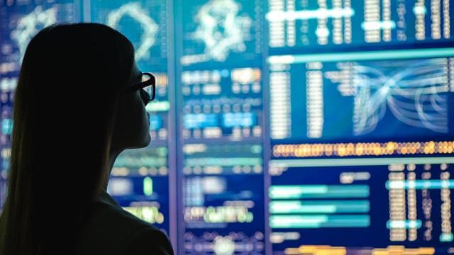 Pessoa em silhueta analisando dados em diversas telas digitais exibindo gráficos e tabelas.
