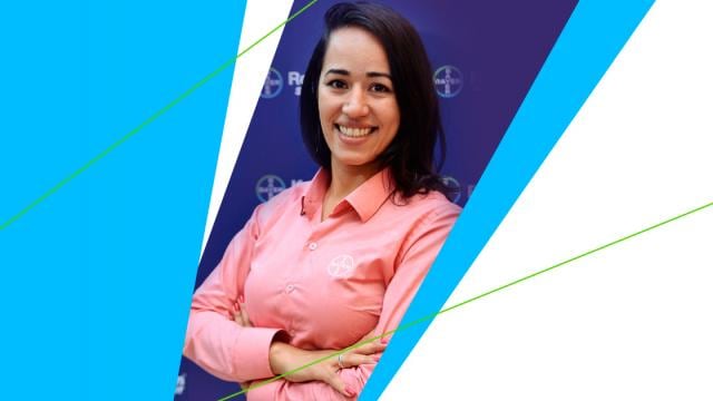 Uma pessoa sorrindo, vestindo uma camisa cor de pêssego com um logotipo, sobre um fundo azul com seções diagonais brancas e azuis.