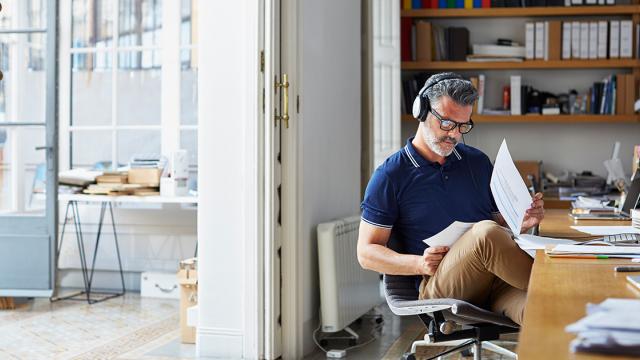 Homem com fones de ouvido sentado em uma mesa, lendo documentos em um escritório bem iluminado, com prateleiras e uma porta aberta ao fundo.