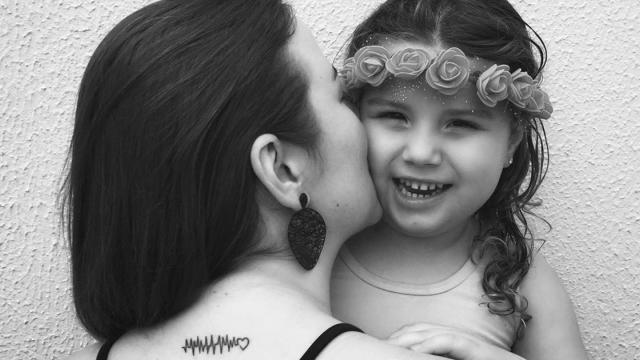 Uma mulher com uma tatuagem de batimento cardíaco beija uma criança sorridente usando uma faixa de flores na cabeça.
