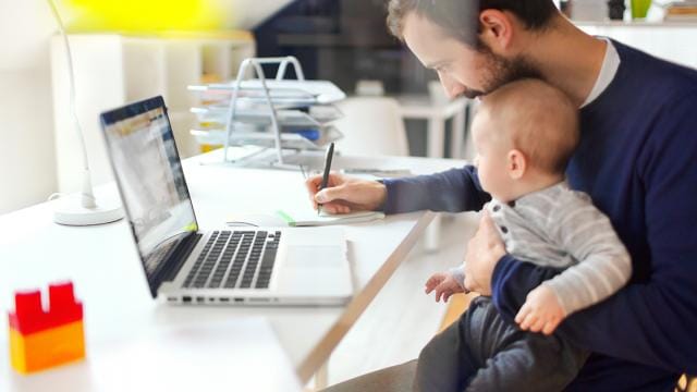 Um homem trabalha em um laptop enquanto segura um bebê no colo.