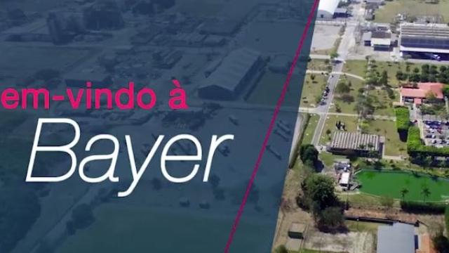Vista aérea mostrando uma unidade da Bayer à direita e a frase "Bem-vindo à Bayer" à esquerda, sobre um fundo azul.