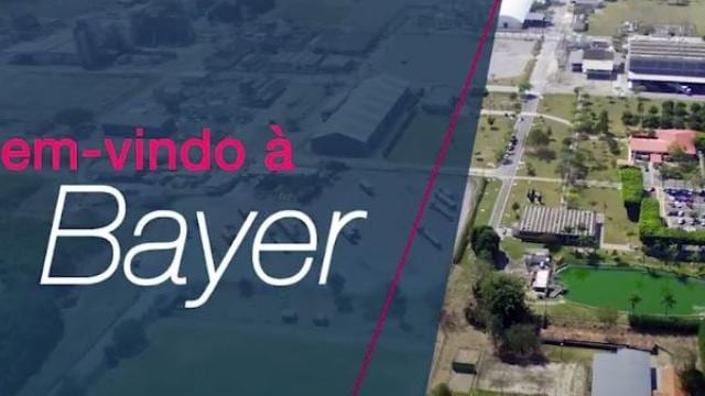 Vista aérea mostrando uma unidade da Bayer à direita e a frase "Bem-vindo à Bayer" à esquerda, sobre um fundo azul.