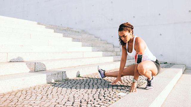 Uma mulher vestida de forma esportiva se alonga em degraus externos, com um smartphone preso ao braço.