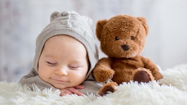 Um bebê vestindo um moletom cinza descansa em uma superfície fofa ao lado de um ursinho de pelúcia marrom.