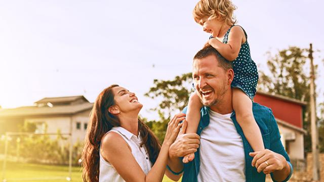 Um homem carrega uma criança nos ombros enquanto uma mulher está ao lado deles, sorrindo ao ar livre em um dia ensolarado.