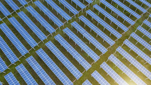 Vista aérea de uma grande fazenda de painéis solares com fileiras de painéis azuis em grama verde.