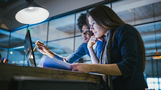 Duas mulheres em um escritório trabalham juntas em um computador, segurando documentos e uma caneta, com uma luz no teto iluminando a mesa.