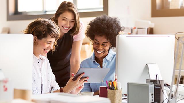 Três pessoas em um ambiente de escritório sorriem enquanto olham para um tablet. Dois desktops e materiais de escritório são visíveis na mesa.