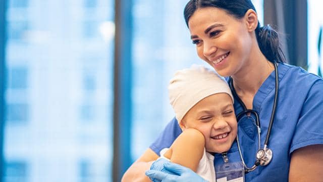 Uma enfermeira de uniforme azul abraça uma criança sorridente usando um lenço na cabeça em um ambiente hospitalar.