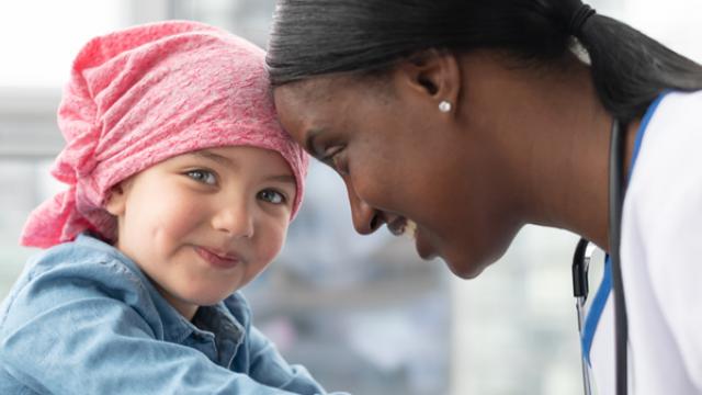 Uma criança sorridente usando um lenço rosa na cabeça está cara a cara com um profissional de saúde de jaleco branco.