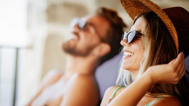 Um homem e uma mulher usando óculos escuros e chapéus estão sentados sorrindo ao sol.