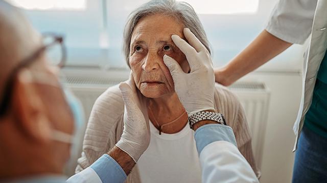 Um médico examina o olho de uma mulher idosa enquanto outra pessoa está por perto em um ambiente médico.