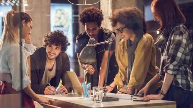 Um grupo de cinco pessoas está reunido ao redor de uma mesa com um laptop, papéis e uma luminária, envolvidos em uma discussão em um ambiente de escritório moderno.