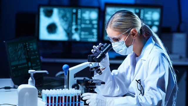 Cientista vestindo jaleco e máscara usa um microscópio, com tubos de ensaio e telas de computador ao fundo.