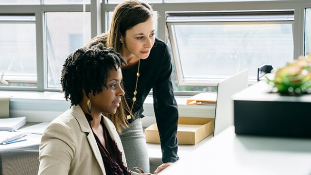 Duas mulheres em um escritório olham juntas para uma tela de computador, uma sentada e a outra em pé atrás dela.