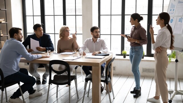Um grupo de pessoas em uma sala de reunião, sentadas e em pé ao redor de uma mesa com papéis e laptops. Duas pessoas estão apresentando ideias perto de um quadro branco.
