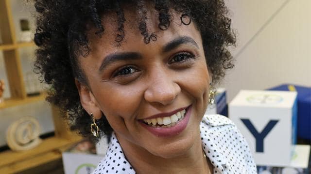 Uma pessoa sorridente com cabelos cacheados e uma camisa de bolinhas está em um ambiente interno perto de prateleiras decoradas.
