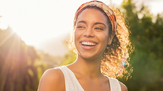 Mulher sorrindo ao ar livre com uma faixa na cabeça, luz do sol iluminando seu rosto e vegetação ao fundo.