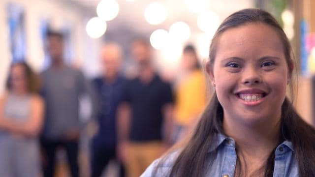 Uma mulher sorrindo em primeiro plano com um grupo de pessoas desfocadas ao fundo.