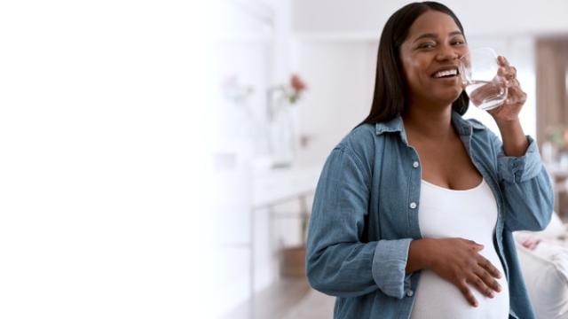 Mulher grávida, de camisa jeans, sorri enquanto bebe água, em pé, dentro de casa.