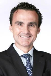 Homem sorridente de terno preto e gravata estampada sobre um fundo simples.