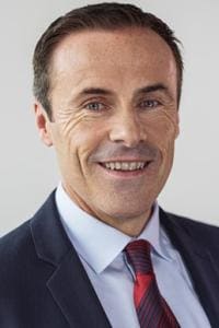 Um homem de terno e gravata sorrindo para a câmera em um fundo neutro.