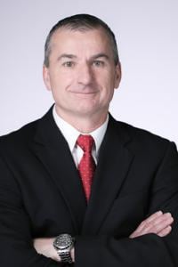 Homem de terno preto com gravata vermelha, braços cruzados e sorrindo. Fundo branco.