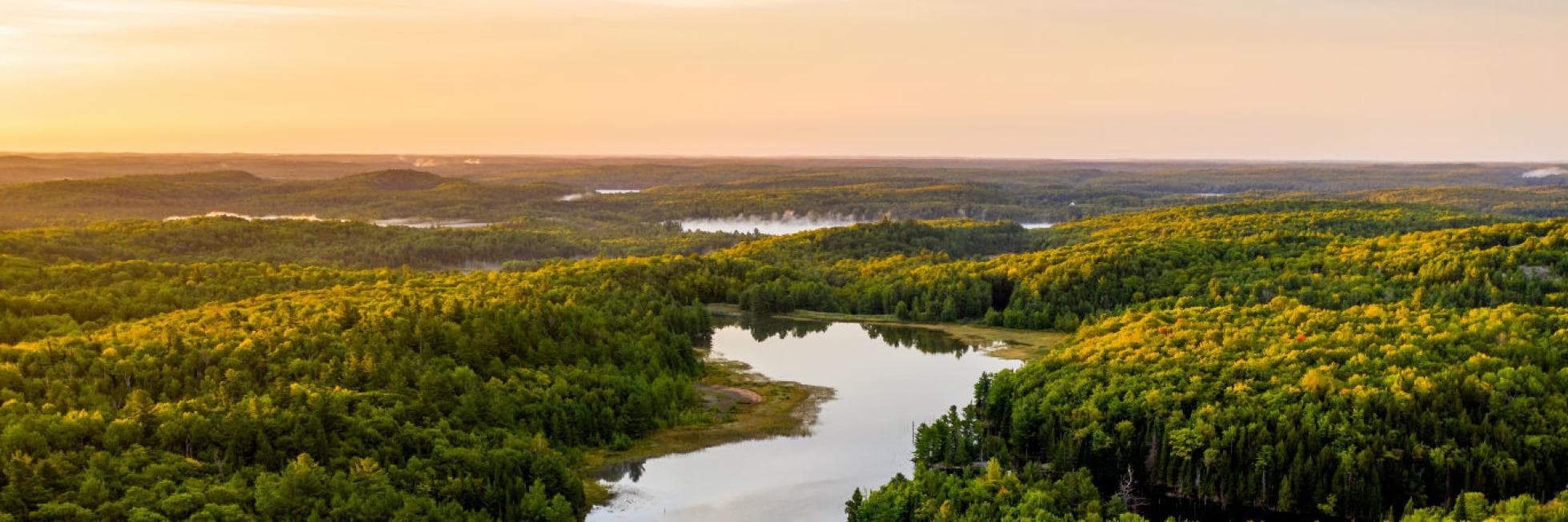 Aerial view of a dense forested landscape with a small lake reflecting the sky, under a warm, golden sunset.